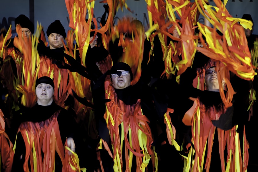 Na zdjęciu teatr ruchu „Balonik”. Uczestnicy ubrani na czarno przyozdobieni czerwono – żółto – pomarańczowymi paskami imitującymi ogień. Tancerze machają rękami.