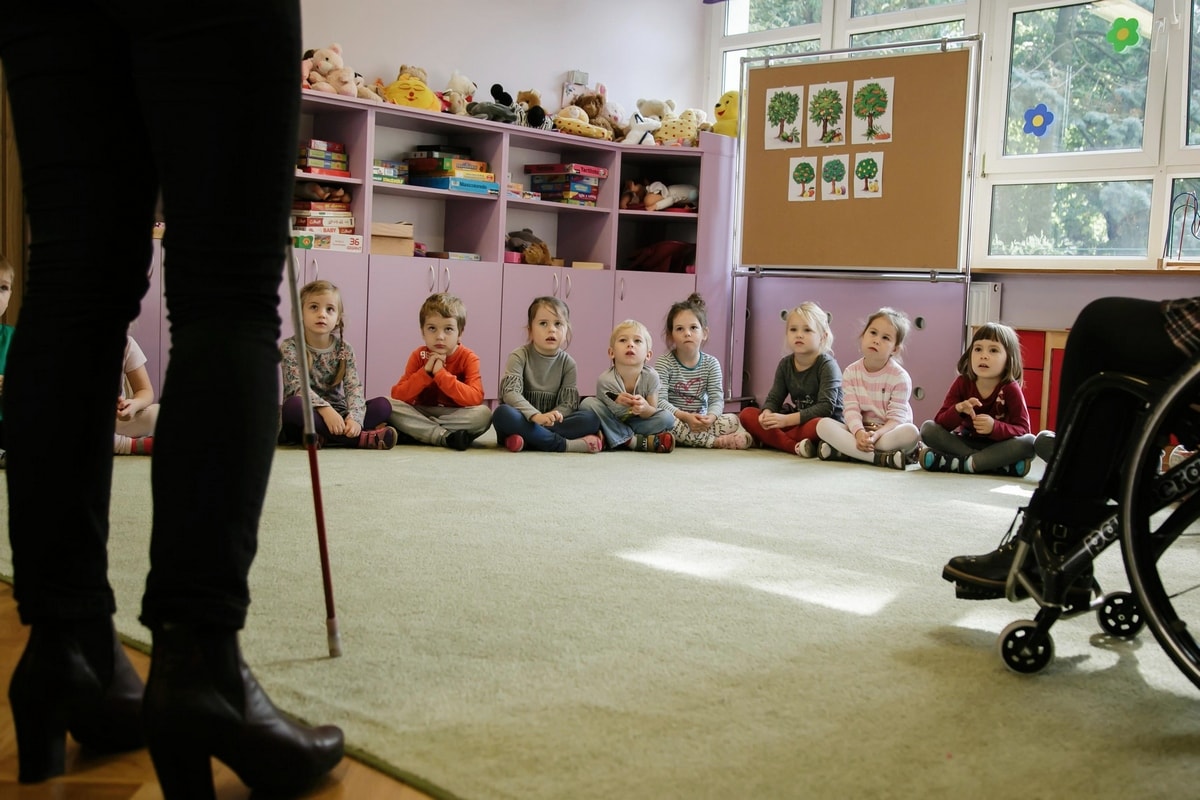 Na zdjęciu sala przedszkolna, w tylnej części sali w półkolu siedzą zasłuchane dzieci. Z lewej strony widać część osoby niewidomej z białą laską , natomiast ze strony prawej wyłania się osoba z niepełnosprawnością ruchu siedząca na wózku.