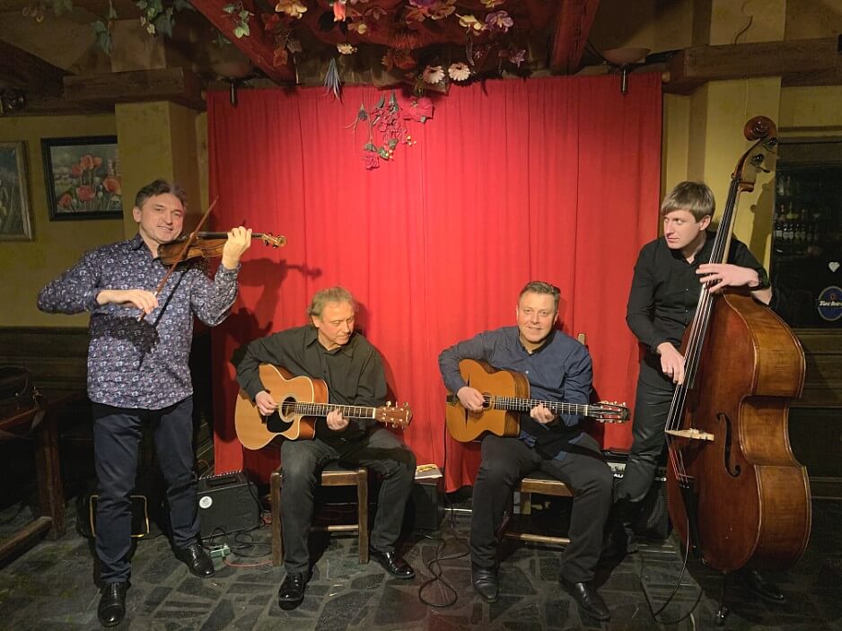 Siergiej Wowkotrub Gypsy Swing Quartet, zdjęcie zespołowe, pierwszy od lewej stojący mężczyzna grający na skrzypcach, dwóch kolejnych mężczyzn siedzących, grających na gitarach, obraz zamyka stojący mężczyzna grający na kontrabasie, ubrany w ciemny garnitur