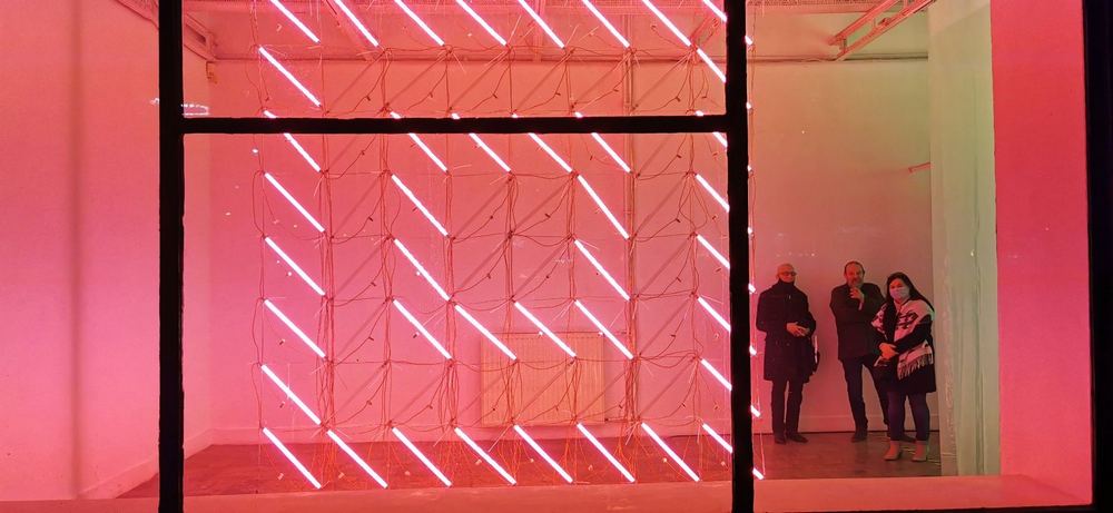 witryna galerii nocą, za szybą widać geometrycznie ułożone świetlówki, świecące na różowo i grupę trzech osób