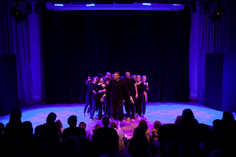 grupa ponad 10 osób w czarnych kostiumach stojących blisko siebie na scenie. na pierwszym planie ciemne sylwetki publiczności