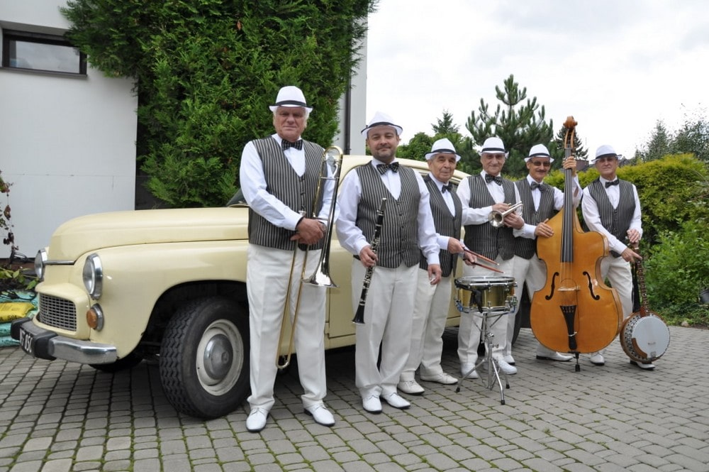 fotografia: sześciu mężczyzn w jednakowych garniturach, trzymają w rękach instrumenty muzyczne, stoją przed starym samochodem marki Warszawa