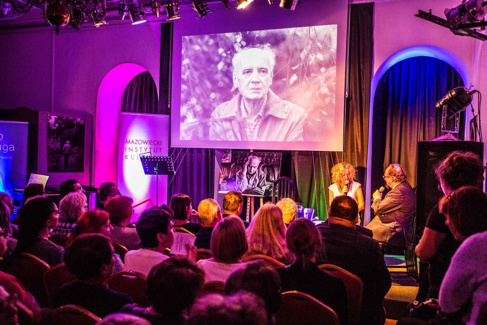 fotografia: publiczność zgromadzona w sali. Na scenie Barbara Gruszka-Zych - poetka i dziennikarka ze Śląska oraz Adam Rozlach - dziennikarz  Polskiego Radia  Widoczny na scenie duży portret Wojciecha Kilara. W klubie nastrojowe światło sceniczne.