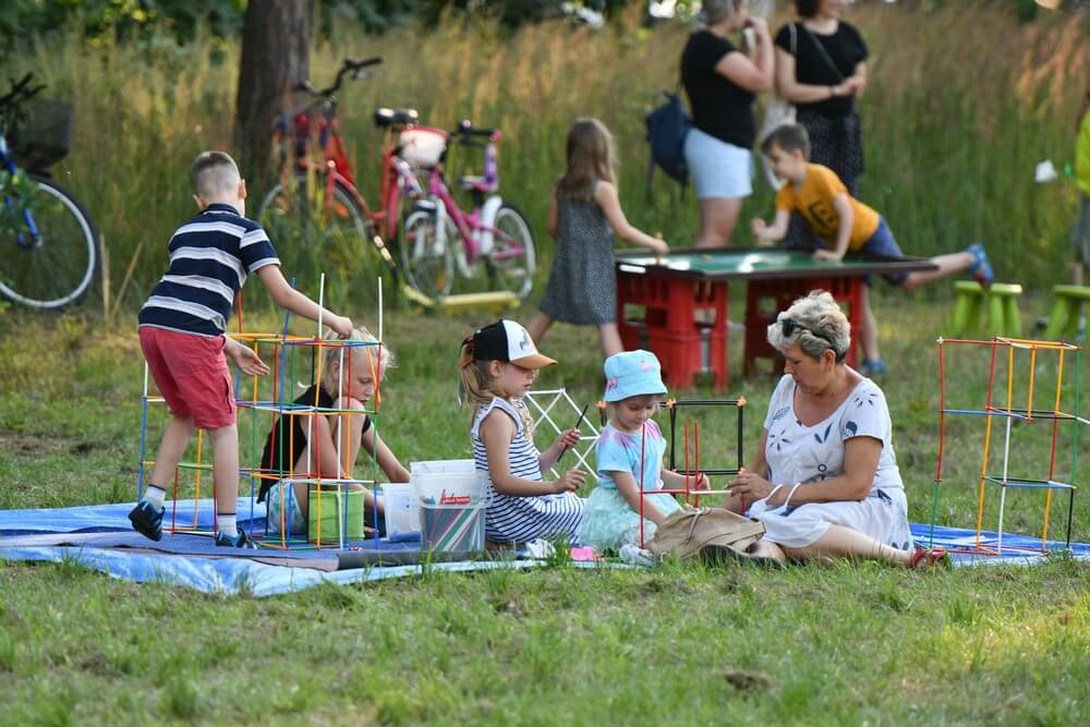 fotografia: dzieci i dorosła kobieta siedzą na trawie i składają konstrukcję z kolorowych rurek