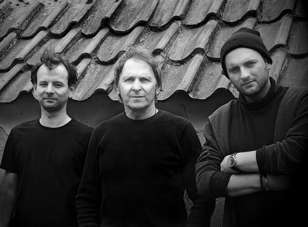 fotografia czarno biała, przedstawia trio artura dutkiewicza: trzech lekko uśmiechających sie mężczyzn, ubranych na ciemno, stojących na tle niskiego budynku, na tle ceramicznej dachówki, stojący po prawej stronie ma dzianinową, czarną czapkę na głowie