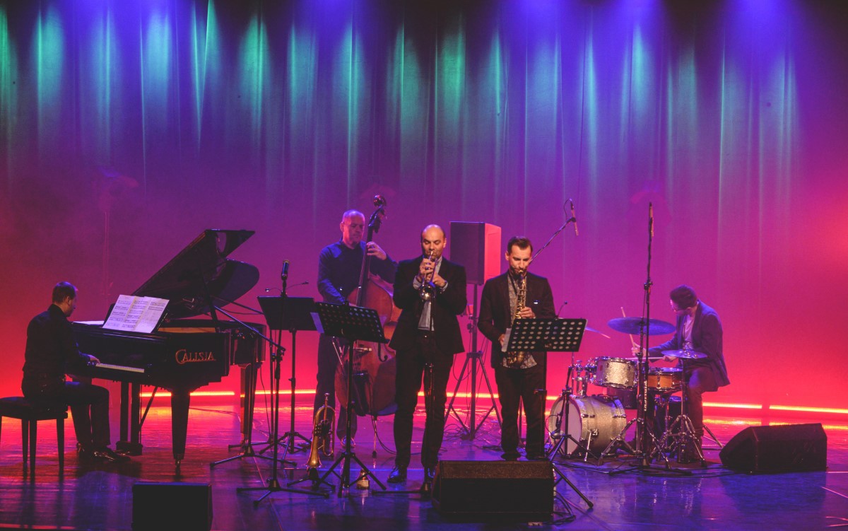 fotografia: pięciu muzyków na scenie, grają na instrumentach, tło podświetlone na czerwono