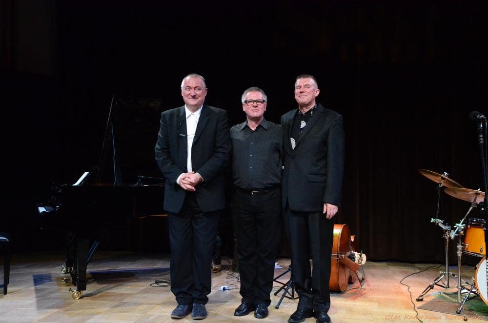 fotografia: trzech pozujących, ubranych na czarno mężczyzn stoi obok siebie na scenie na tle fortepianu, kontrabasu i perkusji
