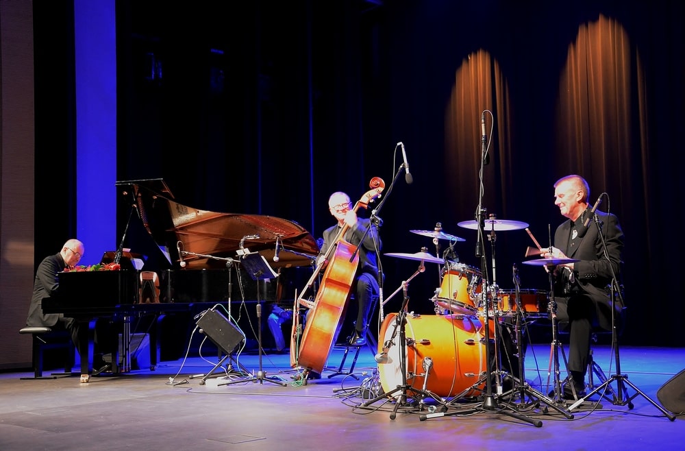 fotografia: zdjęcie sceniczne zespołu, trzech mężczyzn w czarnych garniturach gra na scenie, pierwszy z lewej na fortepianie, kolejny na kontrabasie, następny na perkusji