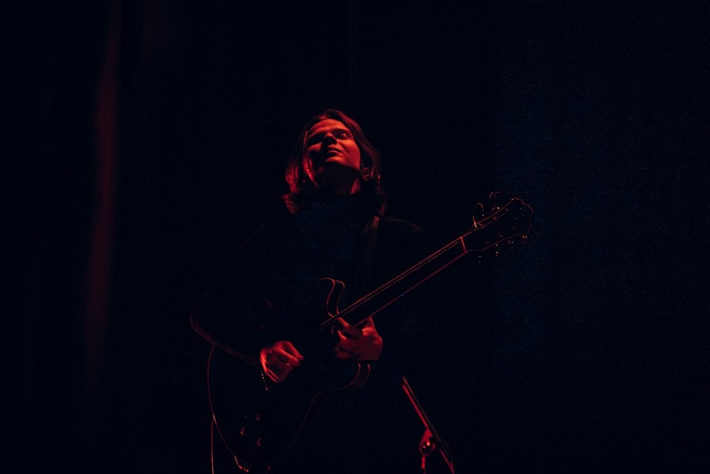 fotografia: na ciemnym tle mało widoczny zarys postaci gitarzysty wyłaniającej się z ciemności