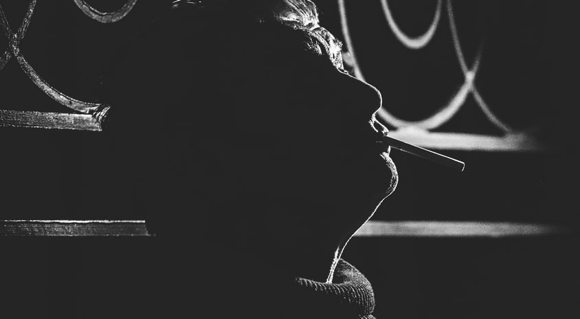 fotografia czarno biała, fragment: marcin świetlicki, ciemne tło, podświetlony zarys głowy, w ustach trzyma papierosa