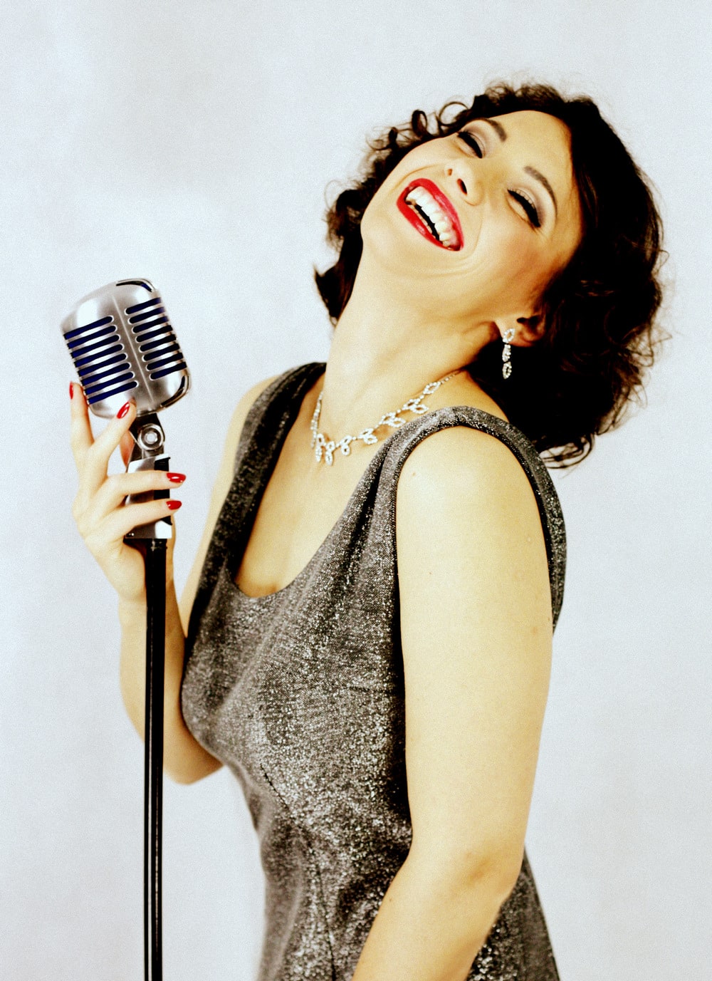 zdjęcie z półprofilu – uśmiechnięta, ciemnowłosa kobieta z odchyloną do tyłu głową, w sukni wieczorowej koloru brązowego, stojąca przy srebrnym mikrofonie na statywie, prawą dłonią dotyka mikrofonu