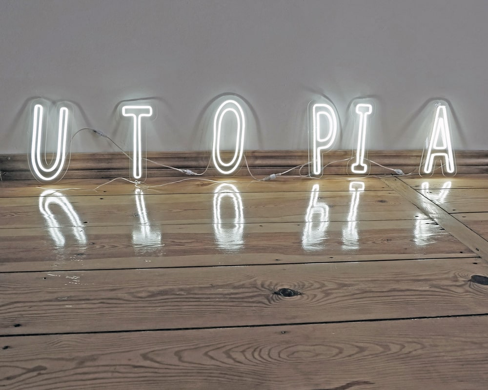 Zdjęcie przedstawia obiekt artystyczny, oparty o ścianę, na drewnianej podłodze. Jest to świecący na biało neon. Neon składa się z pojedynczych liter, tworzących napis: UTOPIA