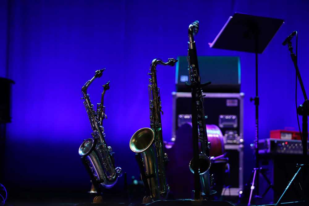 zdjęcie sceniczne w tonacji niebiesko-fioletowej – trzy saksofony. od lewej – sopranowy, altowy, tenorowy, oraz klarnet basowy ustawione na scenie na statywach, w tle kontrabas i sprzęt muzyczny – wzmacniacze