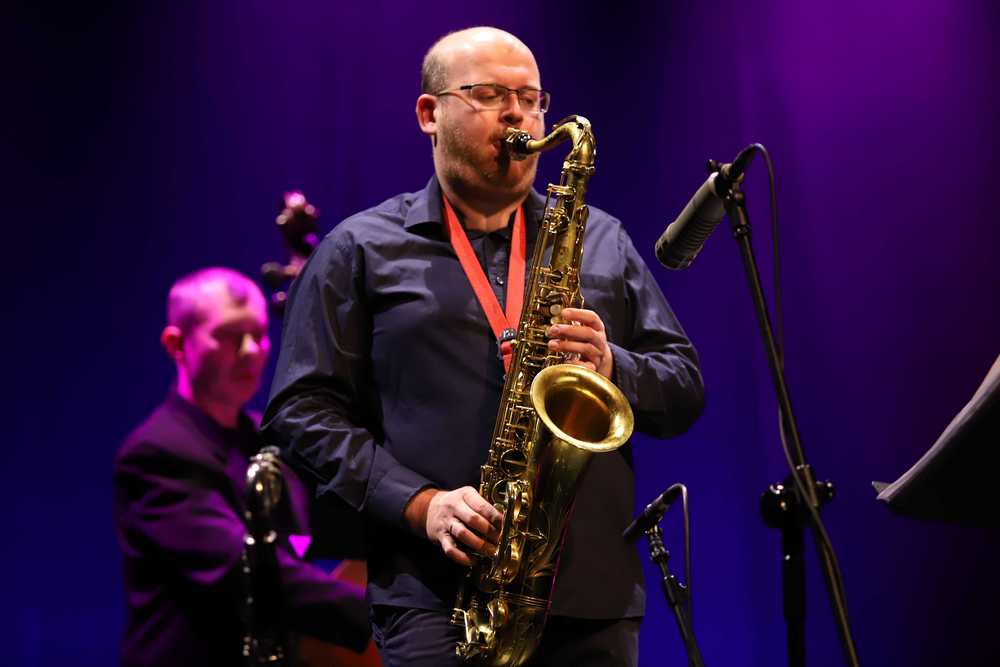 fotografia, zdjęcie sceniczne bliskiego planu, plan tak zwany amerykański – Szymon Łukowski grający na saksofonie tenorowym do mikrofonu na statywie, w tle Wojtek Pulcyn gra na kontrabasie, zdjęcie w tonacji niebiesko-fioletowej