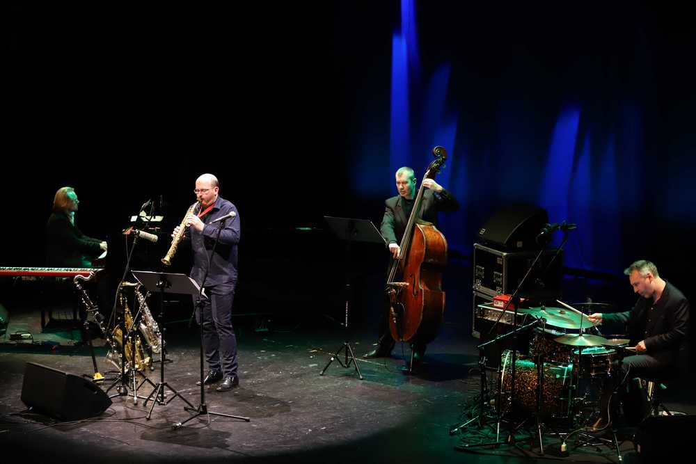 zdjęcie sceniczne w tonacji czarno-niebieskiej, zespół Szymon Łukowski Quartet – czterech mężczyzn ubranych w ciemne garnitury, grających na: od lewej – fortepianie, saksofonie sopranowym, kontrabasie, perkusji