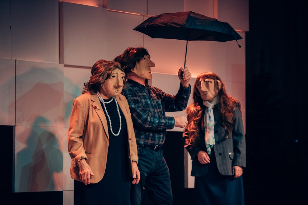 fotografia: troje aktorów w maskach na twarzy, na scenie. jeden z aktorów trzyma w ręce rozłożony parasol