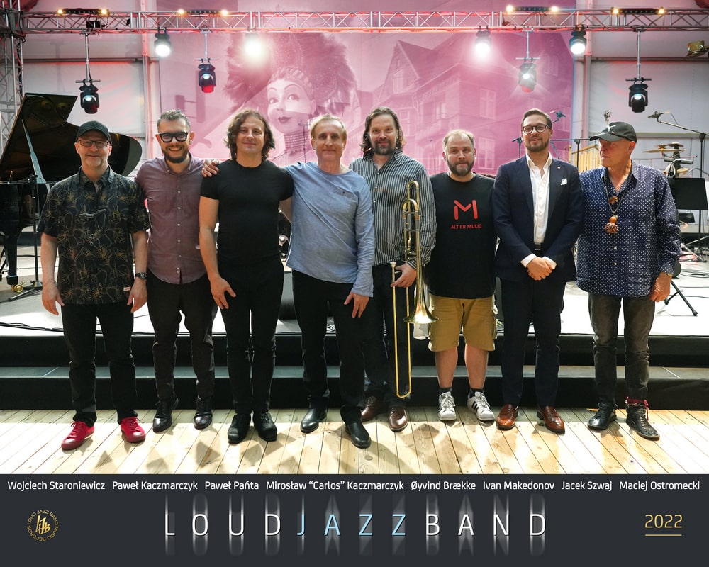 fotografia: zespół Loud Jazz Band_fot. Hubert Gorczyca (zdj. w kolorze, ośmiu uśmiechniętych mężczyzn ustawionych w jednym rzędzie (piąty od lewej trzyma w ręku puzon) pozuje na tle sceny koncertowej, w tle widać fortepian po lewej stronie, z prawej perkusję, z tyłu ekran z wizualizacjami.