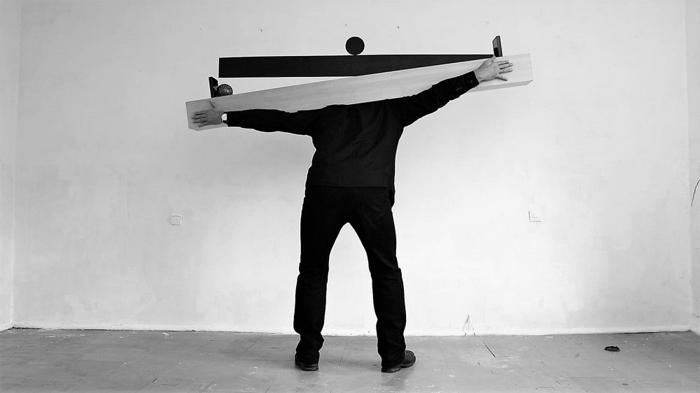 Czarno - białe zdjęcie przedstawia artystę podczas performensu. Mężczyzna stoi przodem do ściany i trzyma na swoich barkach drewnianą belkę