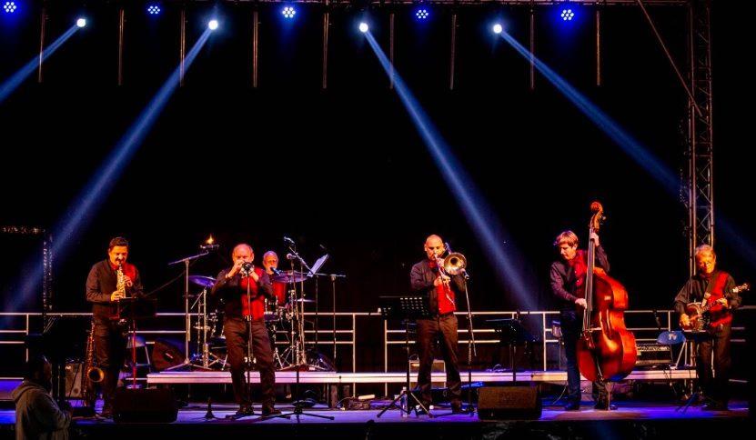 fotografia: sześciu muzyków na scenie gra na instrumentach, z góry oświetleni refletorami