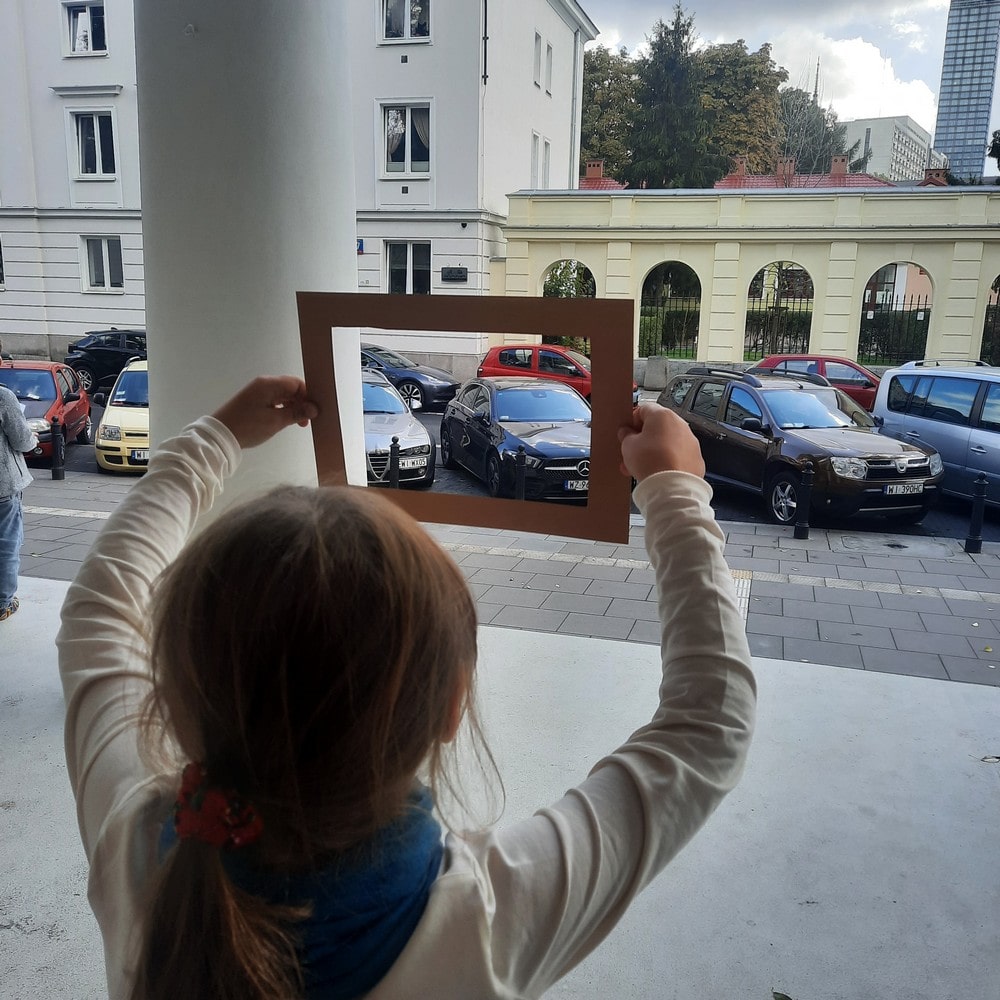 fotografia dziewczynka sfotografowana od tyłu, trzyma w rękach wyciętą ramkę przez którą patrzy na ulicę