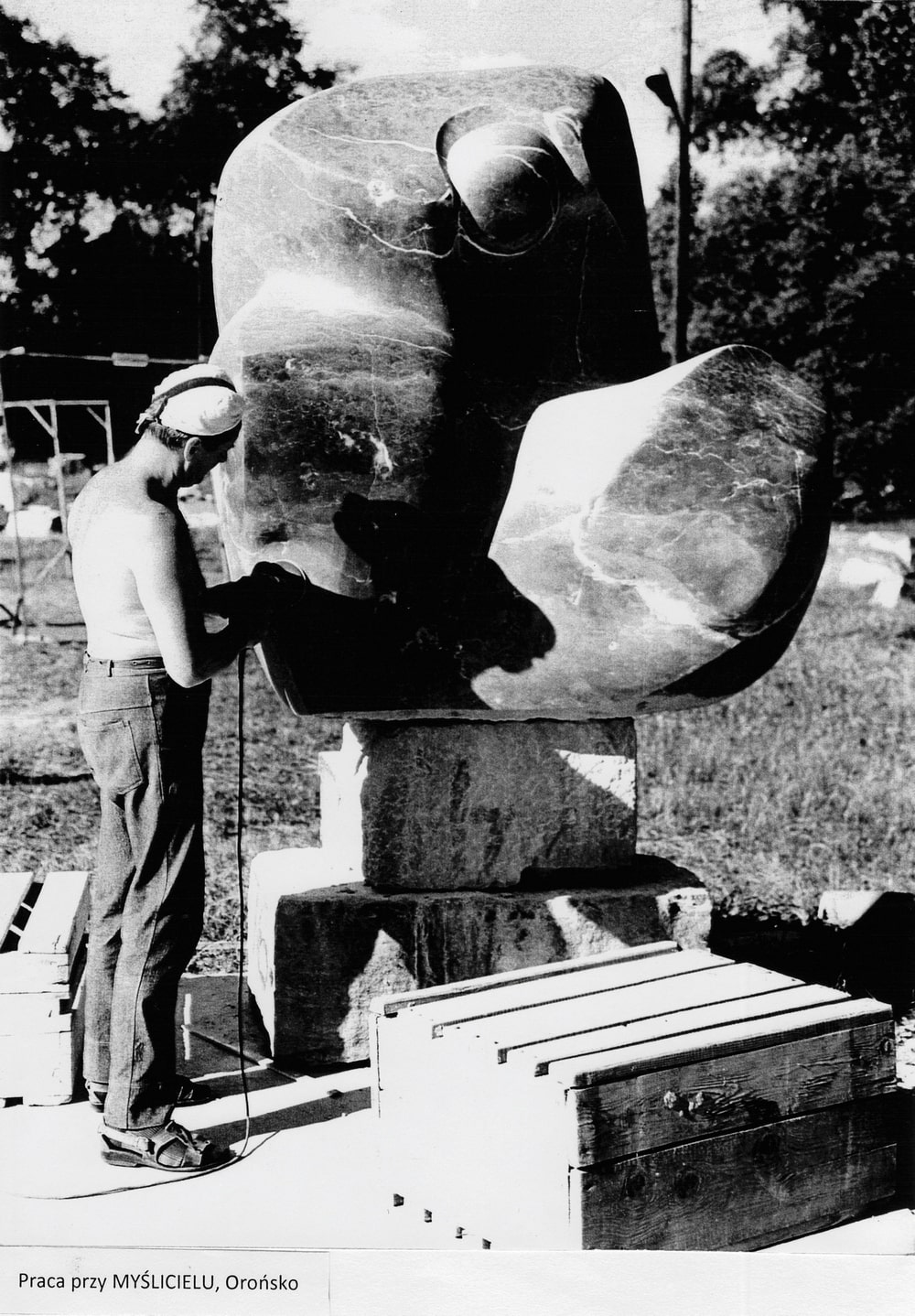 Na zdjęciu widać artystę Kastena podczas pracy nad rzeźbą. Jest ciepło. Artysta pracuje na świeżym powietrzu w plenerze. Rzeźba, którą robi, jest wielkości dorosłego człowieka. Rzeźba przedstawia postać, która siedzi, a głowę ma pochyloną. Postać ma syntetyczne kształty. Zdjęcie jest czarno-białe.