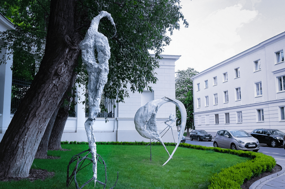 fotografia trzy rzeźby abstrakcyjne na trawniku na tle białych budynków i samochodów stojących na ulicy