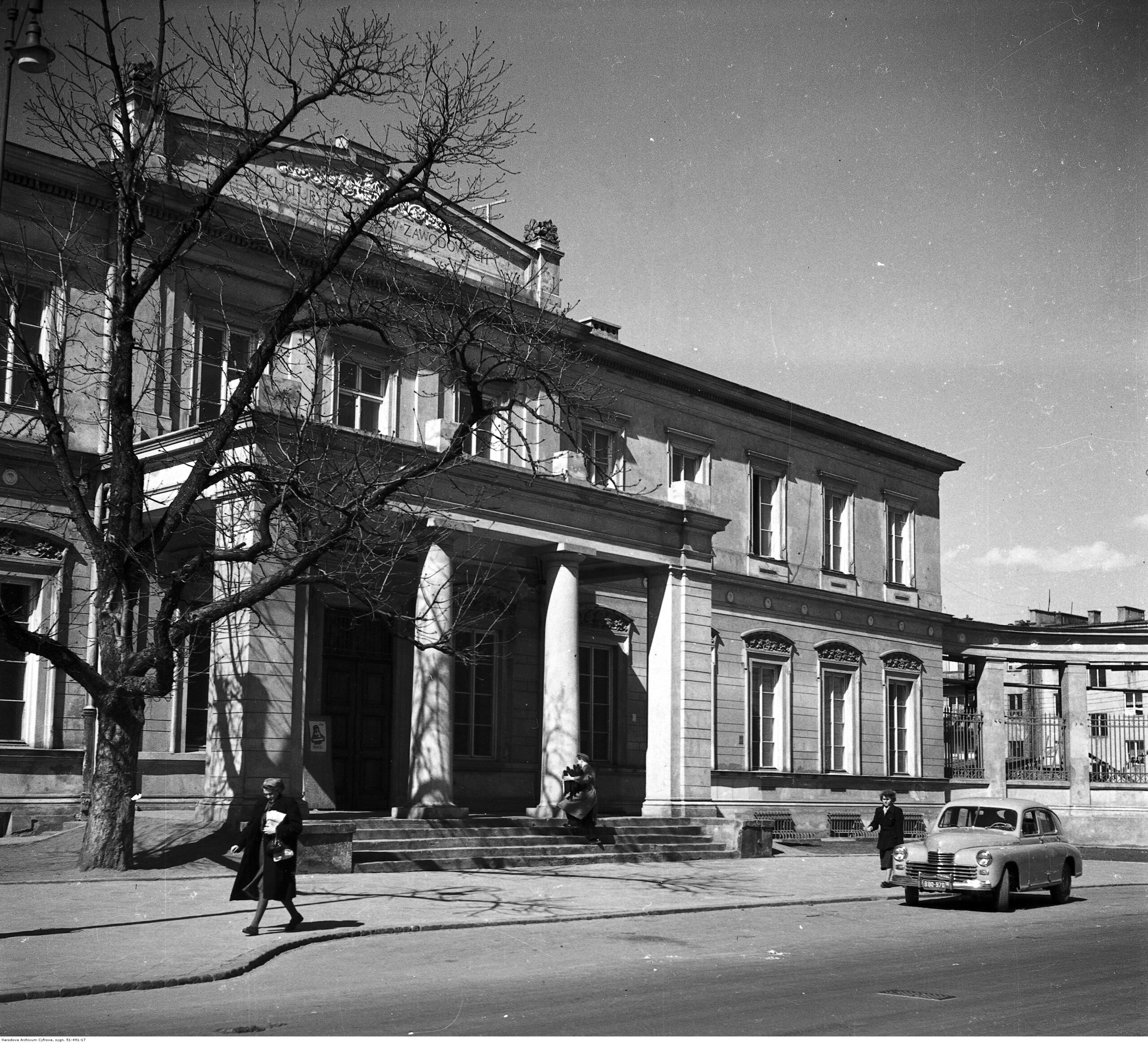 Zdjęcie przedstawiające budynek MIK, na chodniku kobieta, na jezdni stoi samochód. 