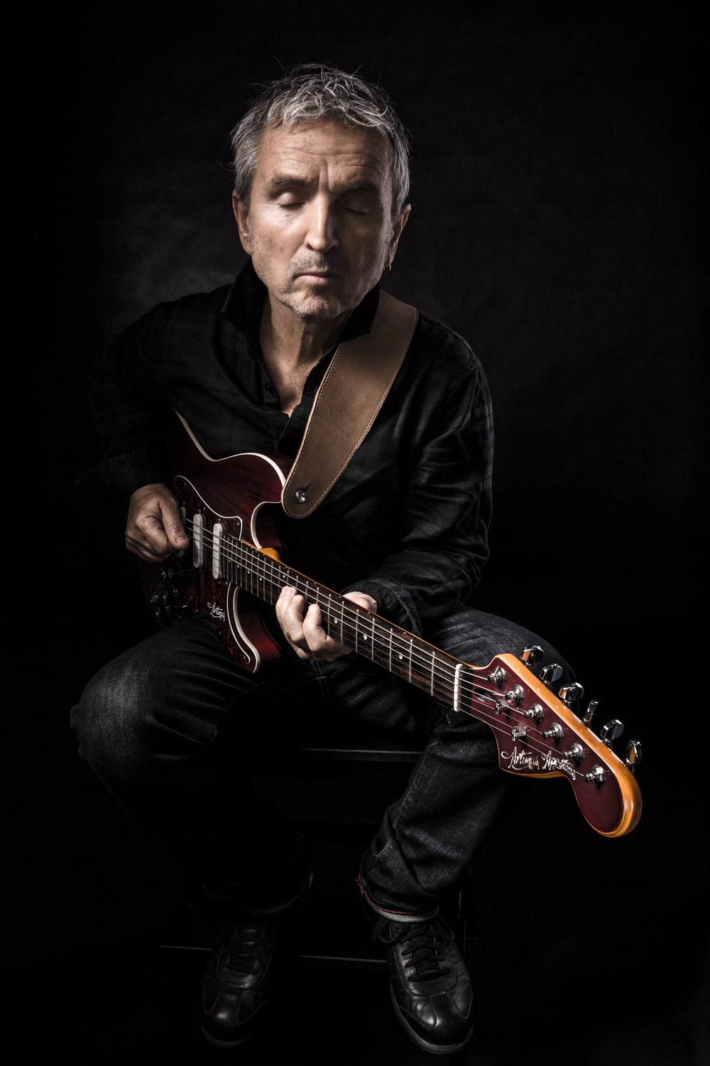 fotografia na ciemnym tle, mężczyzna w pozycji siedzącej, grający na gitarze elektrycznej, ubrany w czarną koszulę i spodnie tegoż koloru