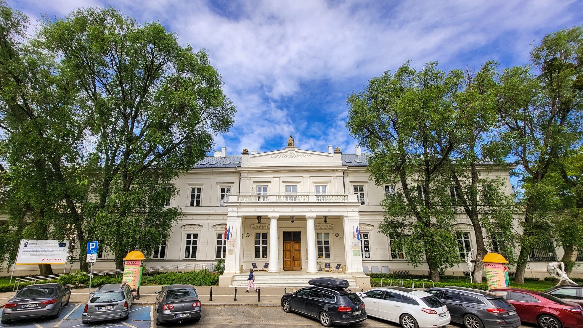 Zdjęcie Mazowieckiego Instytutu Kultury. Budynek przy Elektoralnej 12, ciepłe kolory widoczni przechodnie, samochody i leżaki przed wejściem/